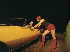 Young woman – perhaps a prostitute – is speaking with a young man who is in his vehicle.