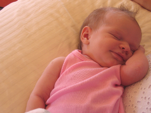 A smiling female baby in a pink outfit rests on a pillow, eyes closed.