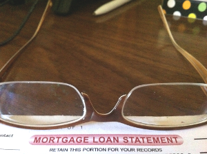 A mortgage loan refinance document sits atop a desk. A pair of eyeglasses rest on top of the document.