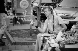A man sits curbside, blowing bubbles with a bubble gun.