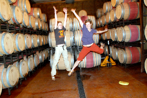 Two young women jump for joy before barrels at a winery.