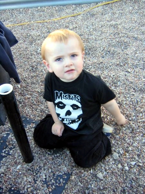 A male toddler is at the playground, wearing a black-and-white Misfits band T-shirt.