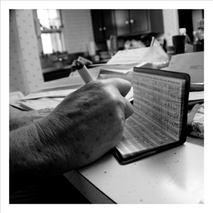 Black-and-white close-up of a checking register. A weathered old hand is writing something in.