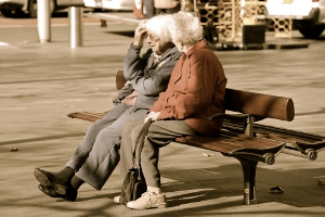 Two gray-haired, elderly women sit together on a wooden bench, reminiscing.
