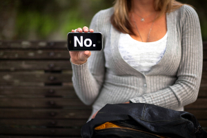 Close-up of a woman seated on a park bench. She's holding a small sign that reads “No.”